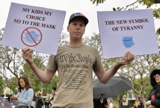 A man wearing a shirt that says 'We the People' holds two signs. One says My Kids My Choice / No to the Mask' and the other says 'The new symbol of tyranny' with an image of a blue face mask