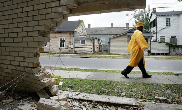 A young person wearing a yellow graduation cap and gown walks down the street, surrounded by wreckage from Hurricane Katrina