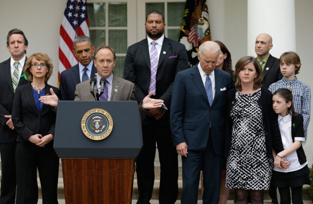 A news photo of Mark Barden standing at a podium outside the White House. Then-President Barack Obama, then-Vice President Joe Biden and others look on