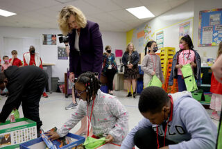 A photo of former Superintendent Vickie Cartwright looking over a student's work in a classroom as other students stand nearby
