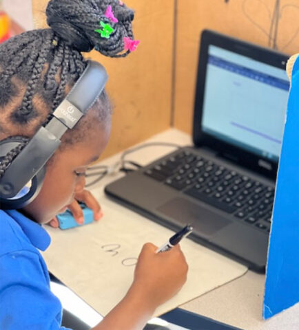 A student wearing headphones writes on paper, laptop open in front of her