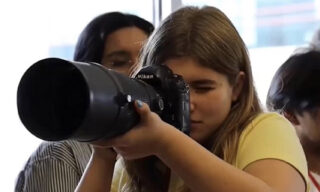 A photo of student journalist Delilah Brumer holding a camera up to her face to shoot a photo