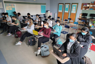 A classroom with about 15 students wearing masks and working on laptops