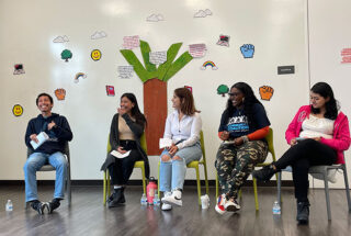 Five young adults sit on chairs during a panel. Student artwork hangs in the background.